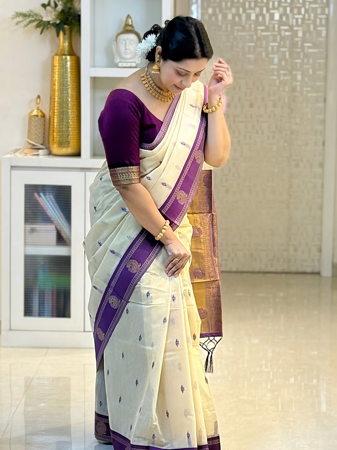 Woman wearing a white and purple saree with gold jewelry in a room setting.