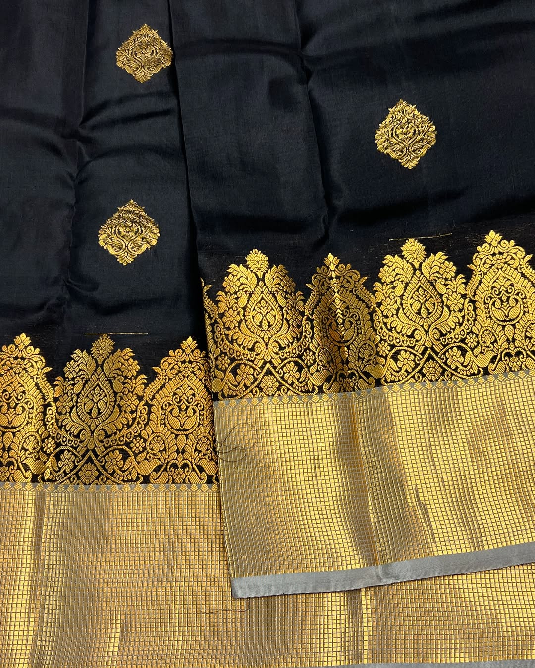 Classic jet black Banarasi Silk saree with scattered gold zari motifs, a wide intricately woven gold zari border, and a shimmering gold/silver zari contrast pallu, elegantly draped on a white textured surface.