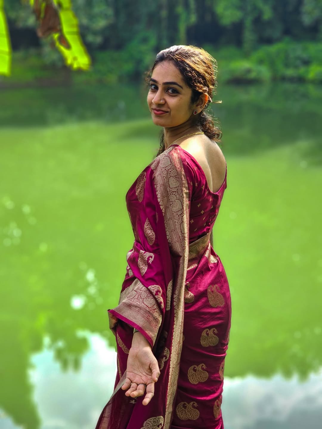 Woman in a pink saree standing by a body of water with greenery in the background