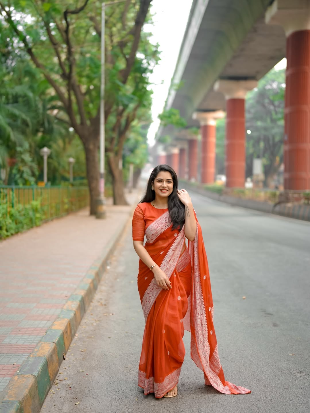 Woman in a red saree standing on a road with greenery and a bridge in the background