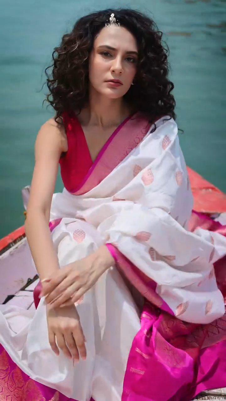 Woman in a white and pink saree sitting on a boat with water in the background