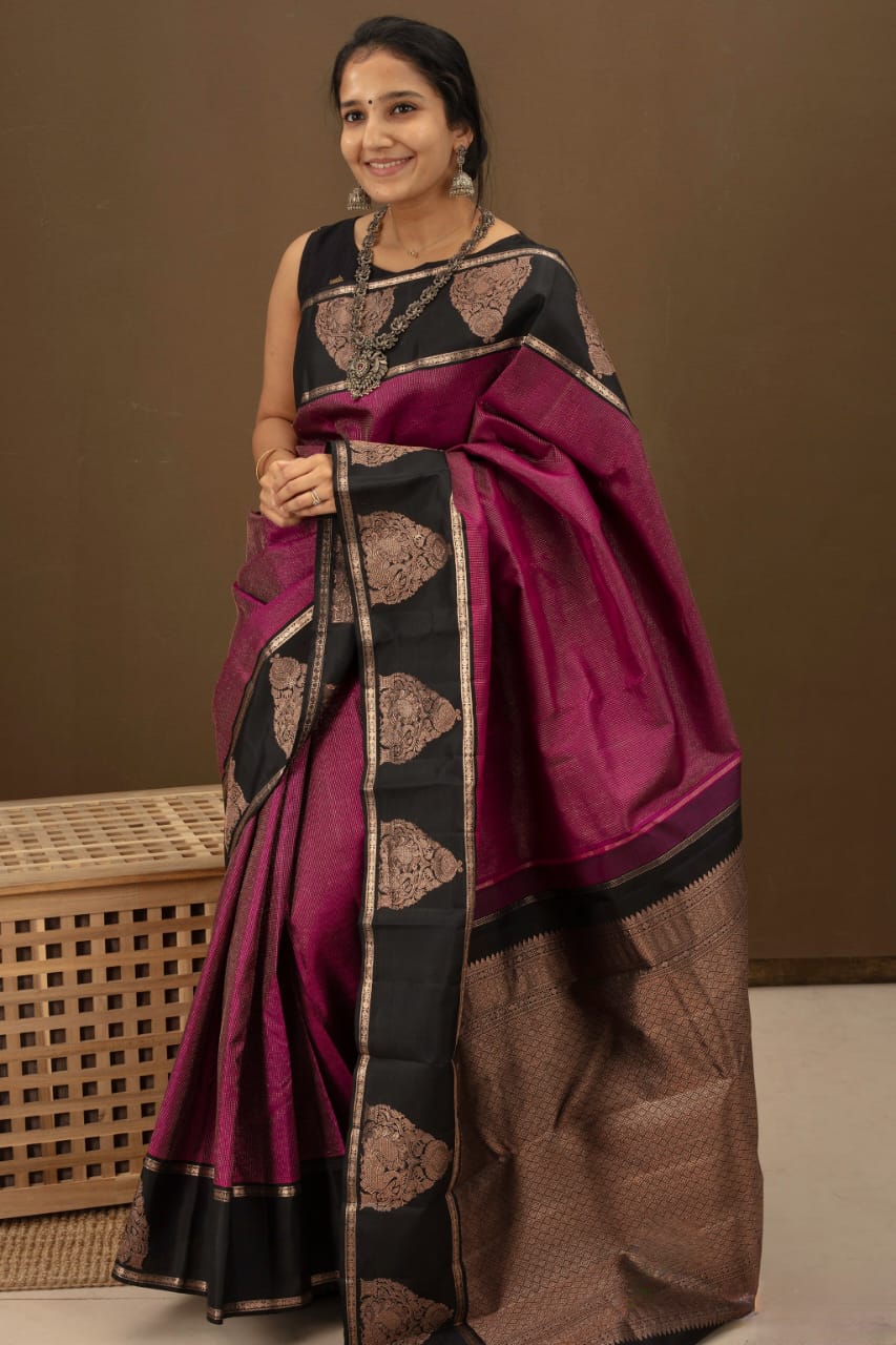 Deep fuschia/magenta Banarasi Silk saree with black contrast panels on the pallu and border, adorned with bronze zari paisley motifs, draped on a woman sitting on a wooden stool indoors.