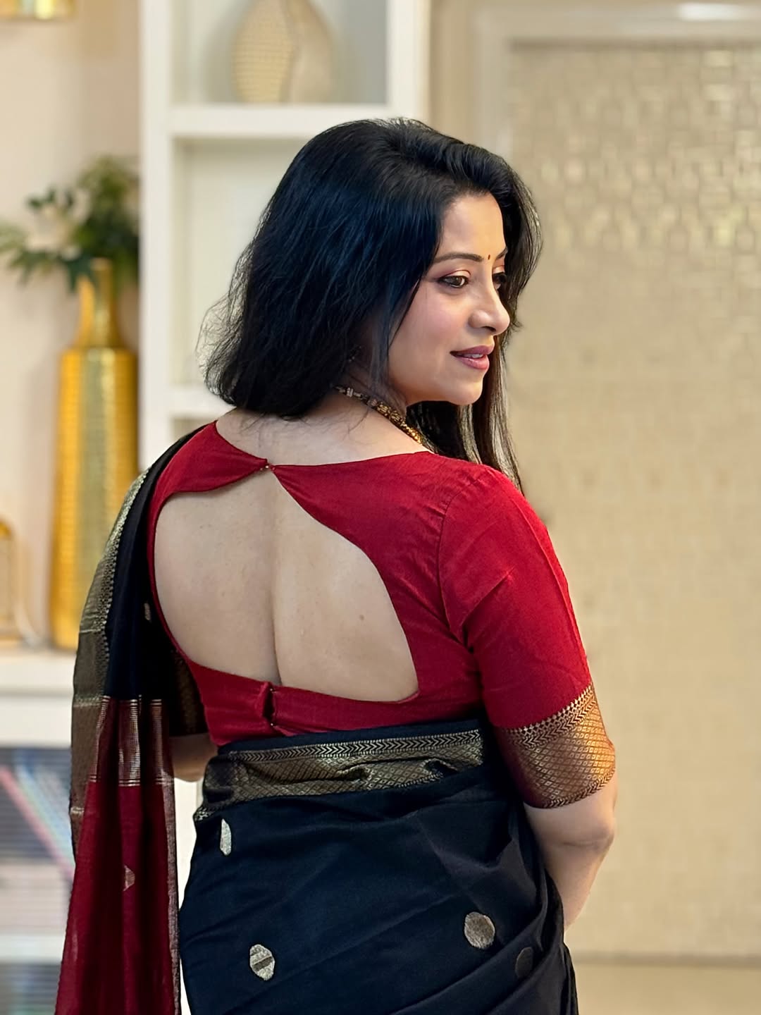 Woman wearing a red blouse with open back and black saree in an indoor setting