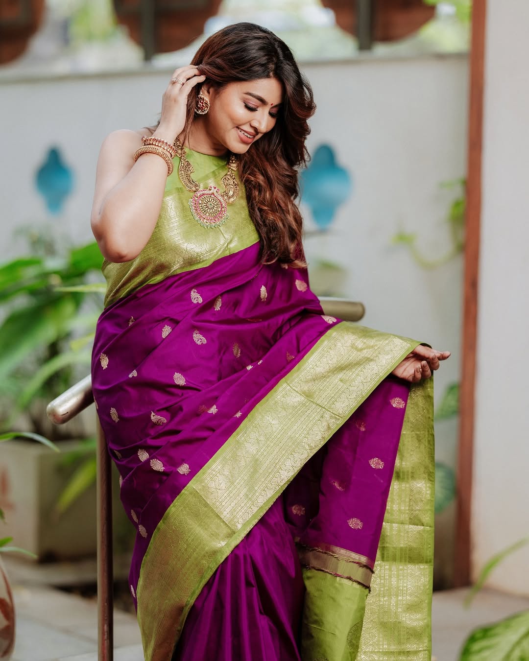 Woman wearing a purple and green saree sitting on a chair with plants in the background
