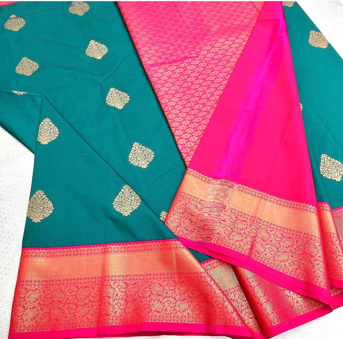 Teal/Peacock Green Banarasi Silk saree with scattered gold zari leaf motifs and a vibrant hot pink and gold zari woven contrast border and pallu, elegantly draped on a white surface.