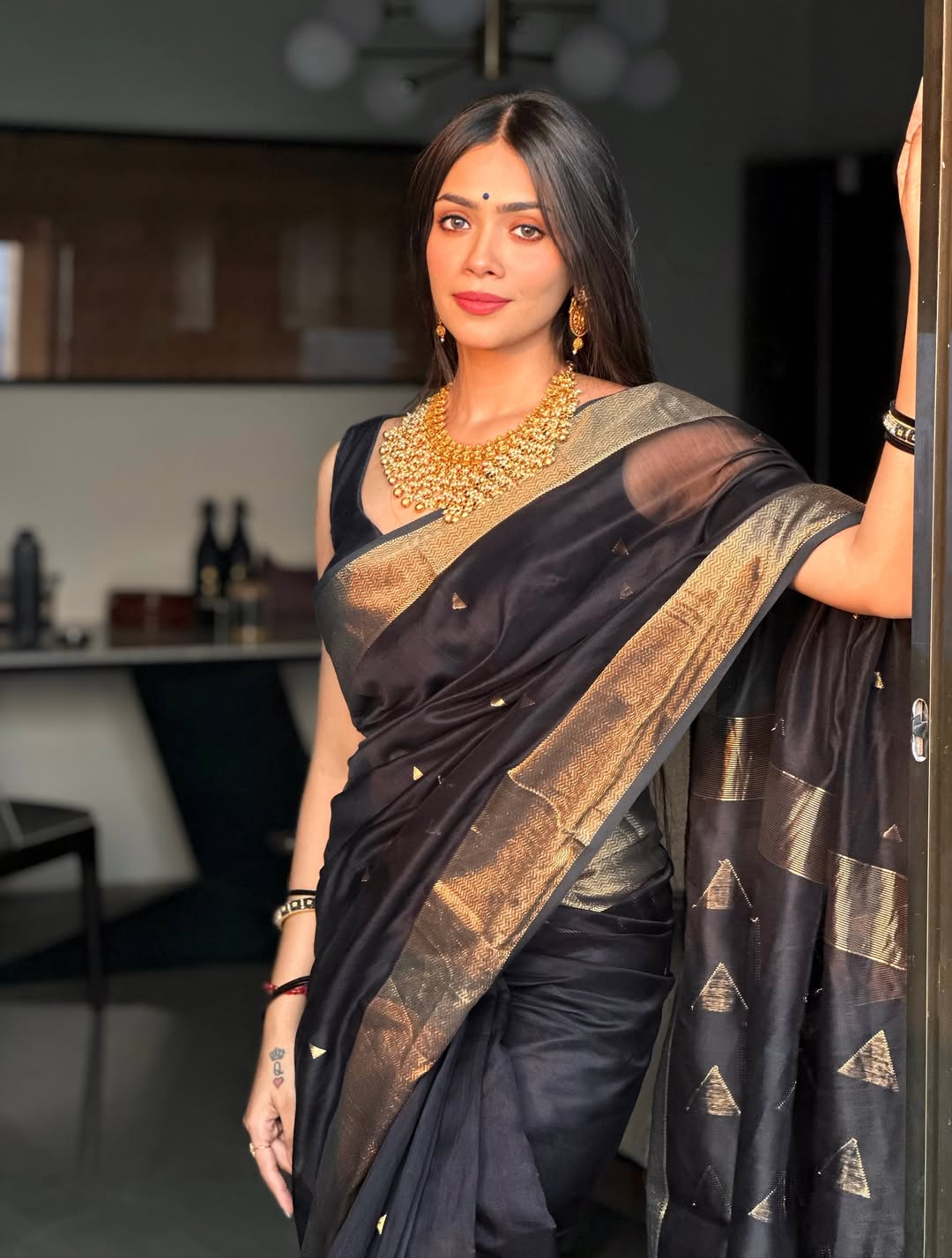 Woman in a black saree with gold patterns standing indoors.