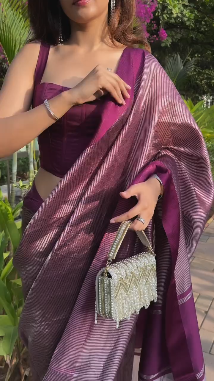 Woman in a purple saree holding a handbag with greenery in the background