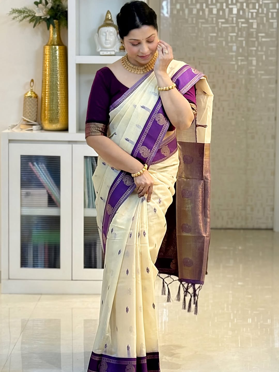 Woman wearing a cream and purple saree in an indoor setting with decorative items.