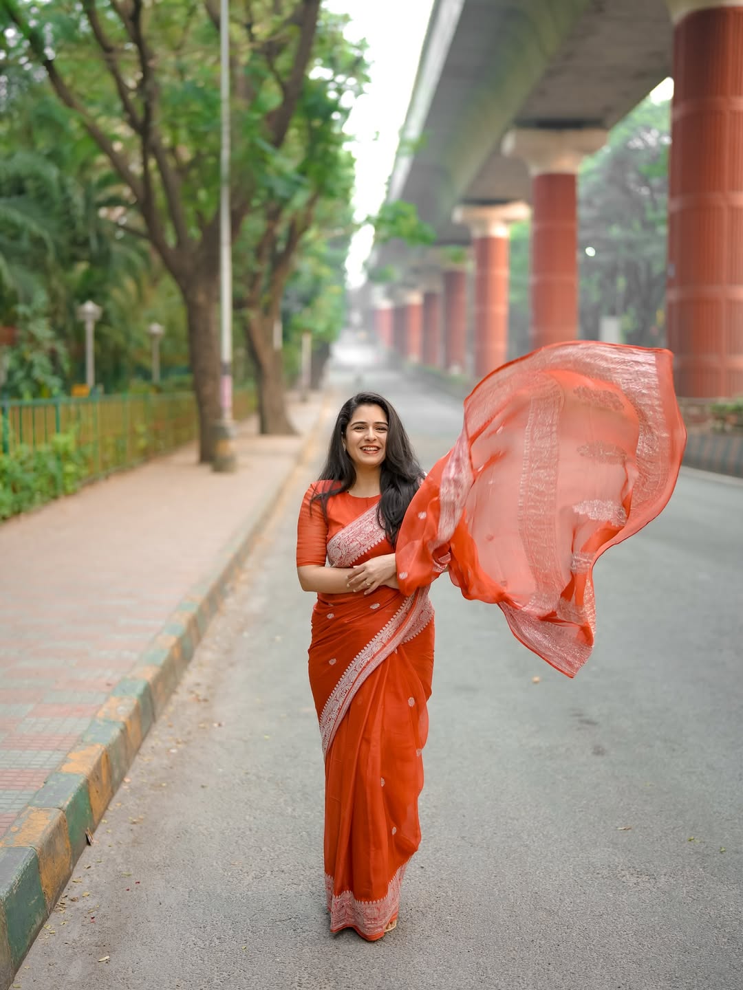 Woman in an orange saree standing on a pathway with trees and a bridge in the background