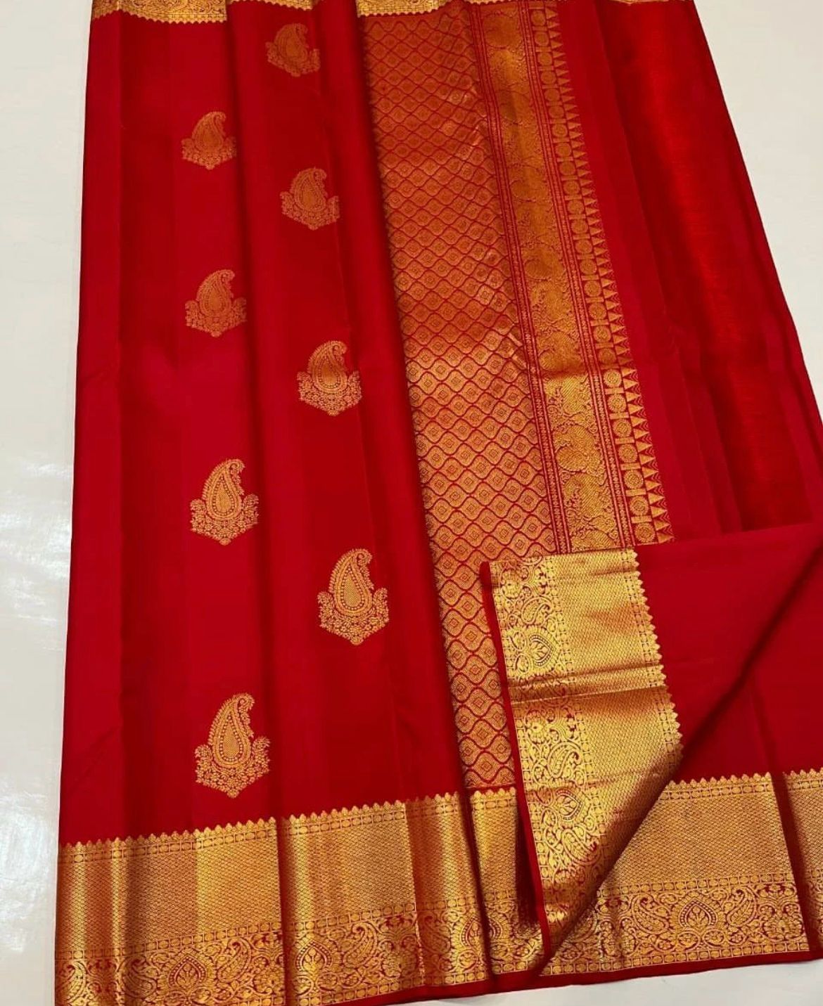 Vibrant rich red Banarasi Silk saree with scattered gold zari mango (paisley) buttas and a wide, intricately woven gold zari border and pallu, elegantly draped on a white surface.