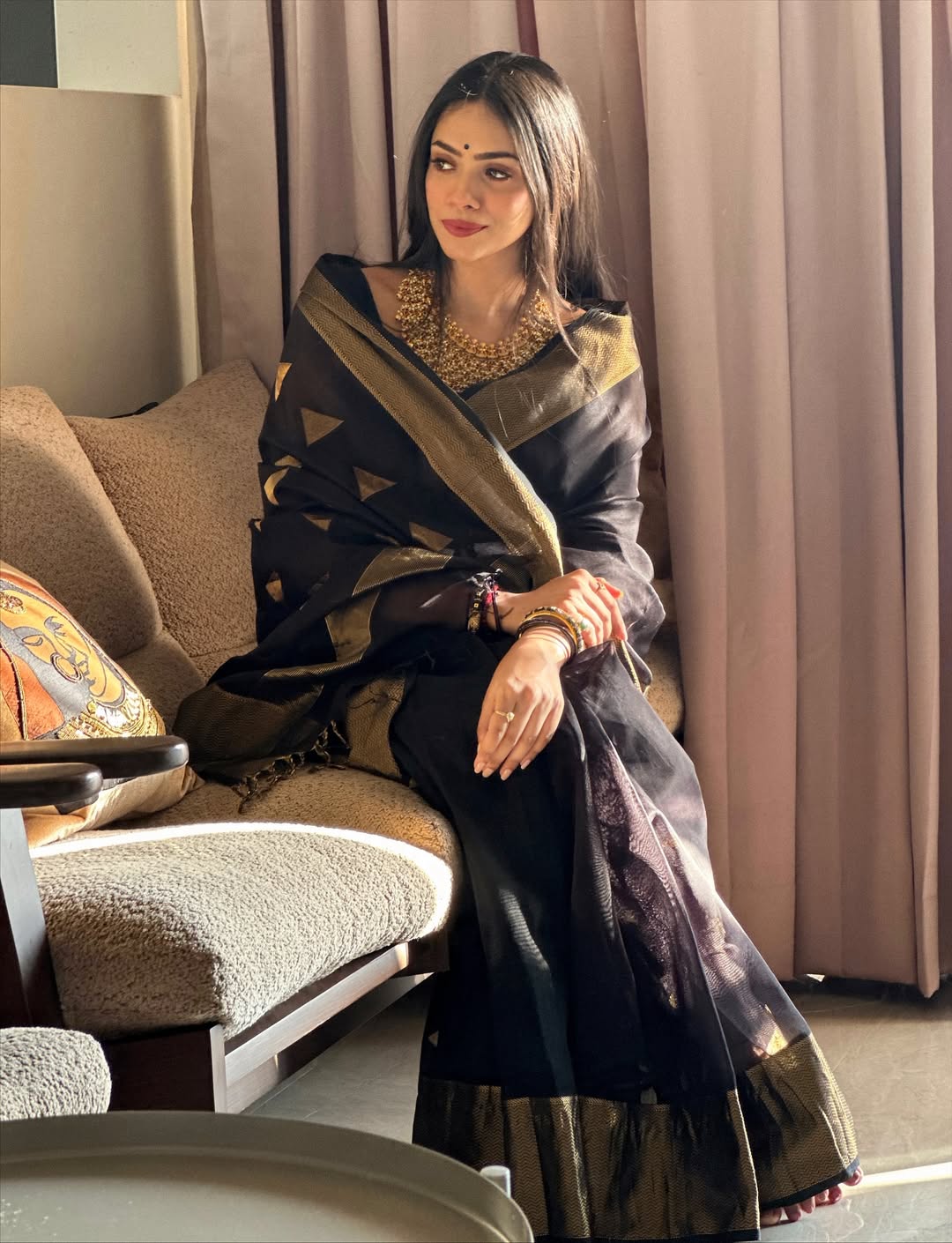 Woman in a black and gold saree sitting on a couch in a room with curtains.