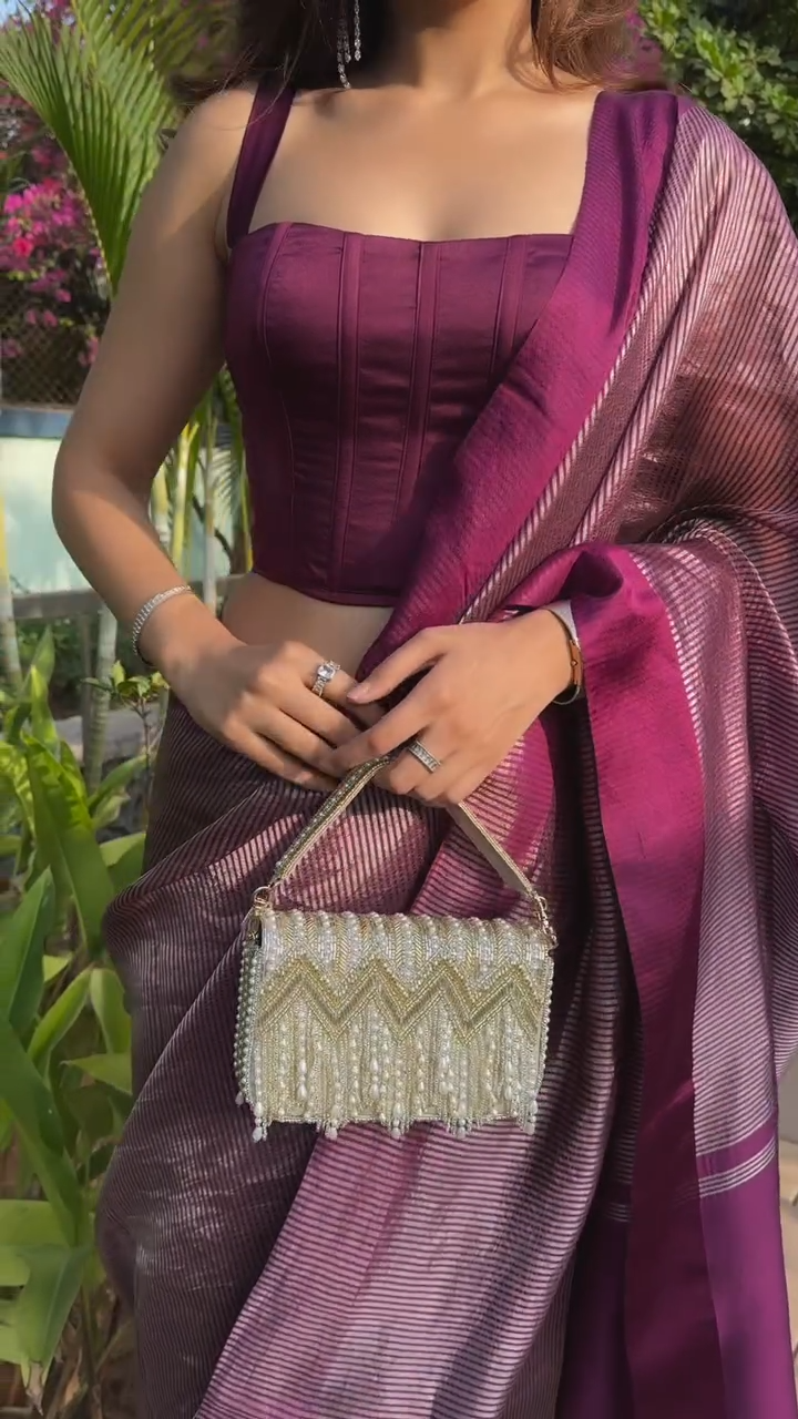 Woman in a purple saree holding a beaded handbag with a garden background