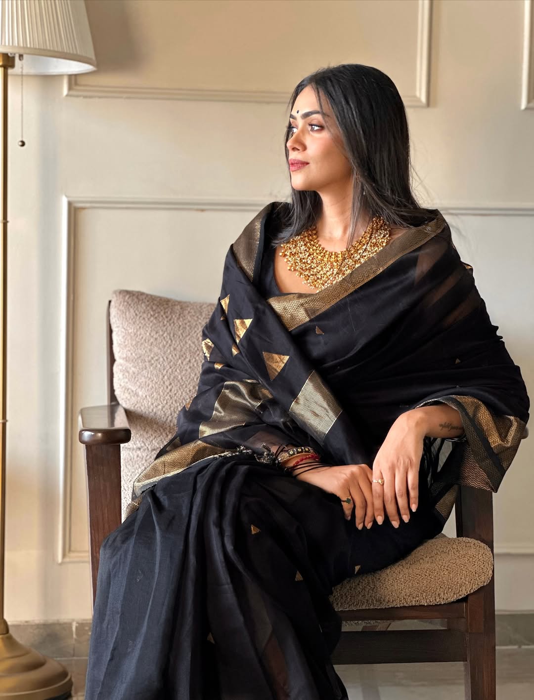 Woman in a black and gold saree sitting on a chair in a room.