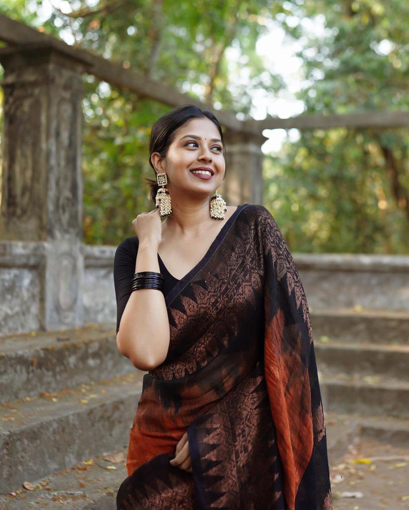 Rust orange Banarasi Silk saree with a black woven border featuring traditional motifs, worn by a smiling woman in an outdoor setting with concrete steps and greenery.