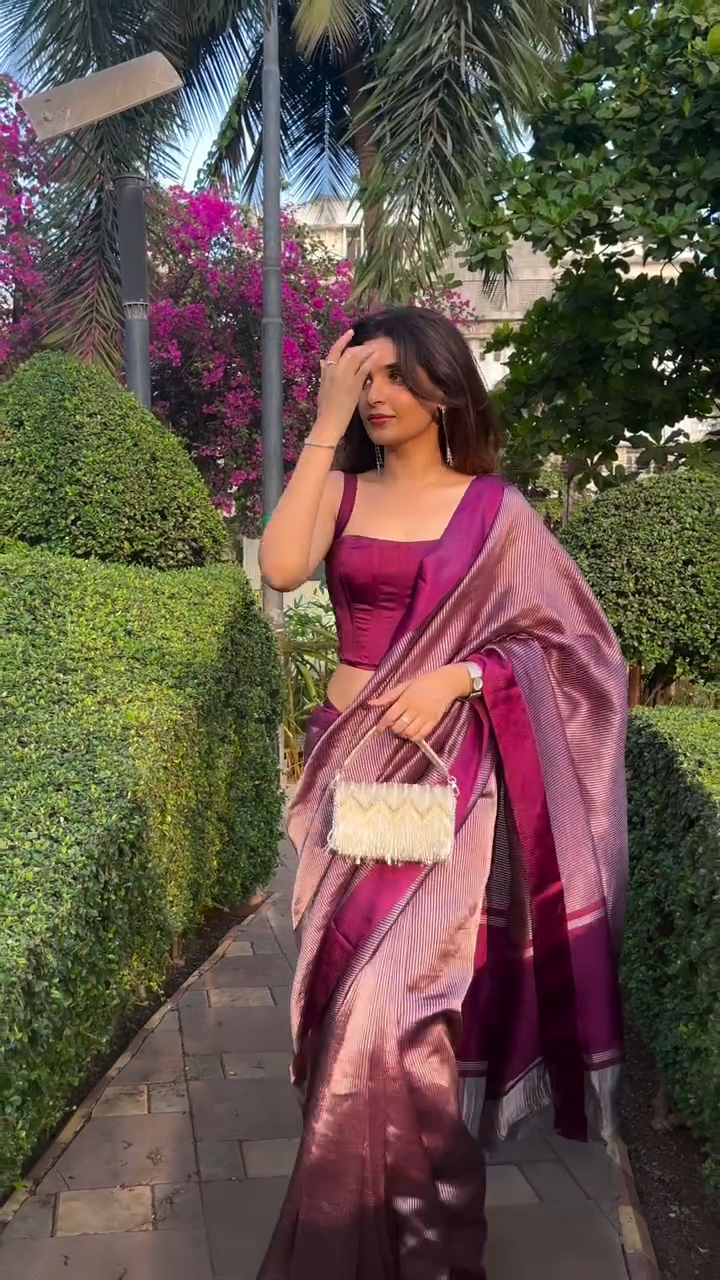 Woman in a purple saree with a matching blouse, standing outdoors with greenery in the background.