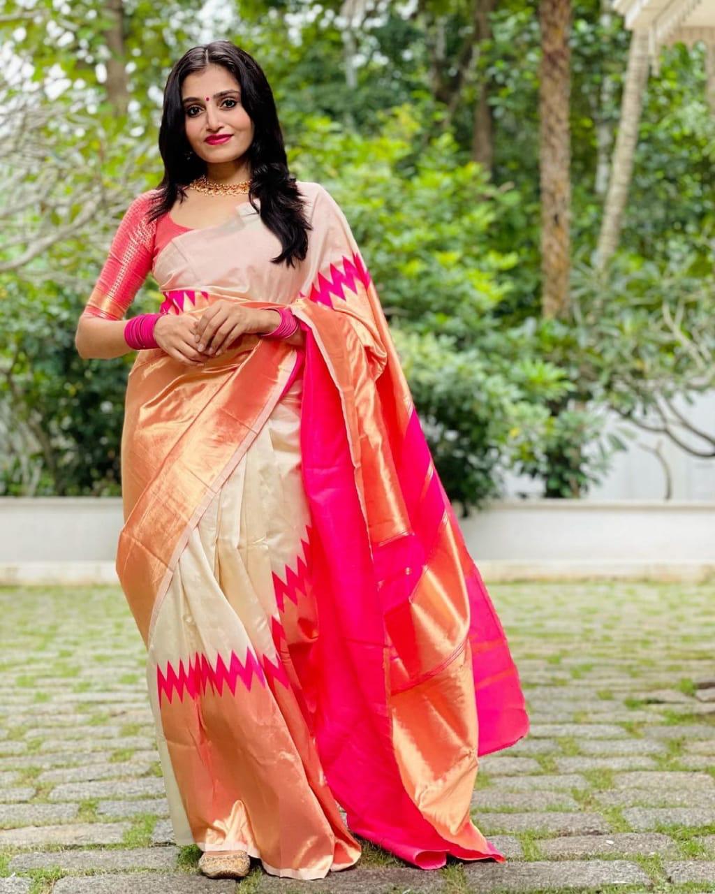 A smiling woman stands against a brick wall, elegantly draped in a soft Lichi silk saree in a blush golden-beige hue, featuring a rich rani pink and golden border, paired with a vibrant red blouse, gold temple jewelry, and gajra in her hair.