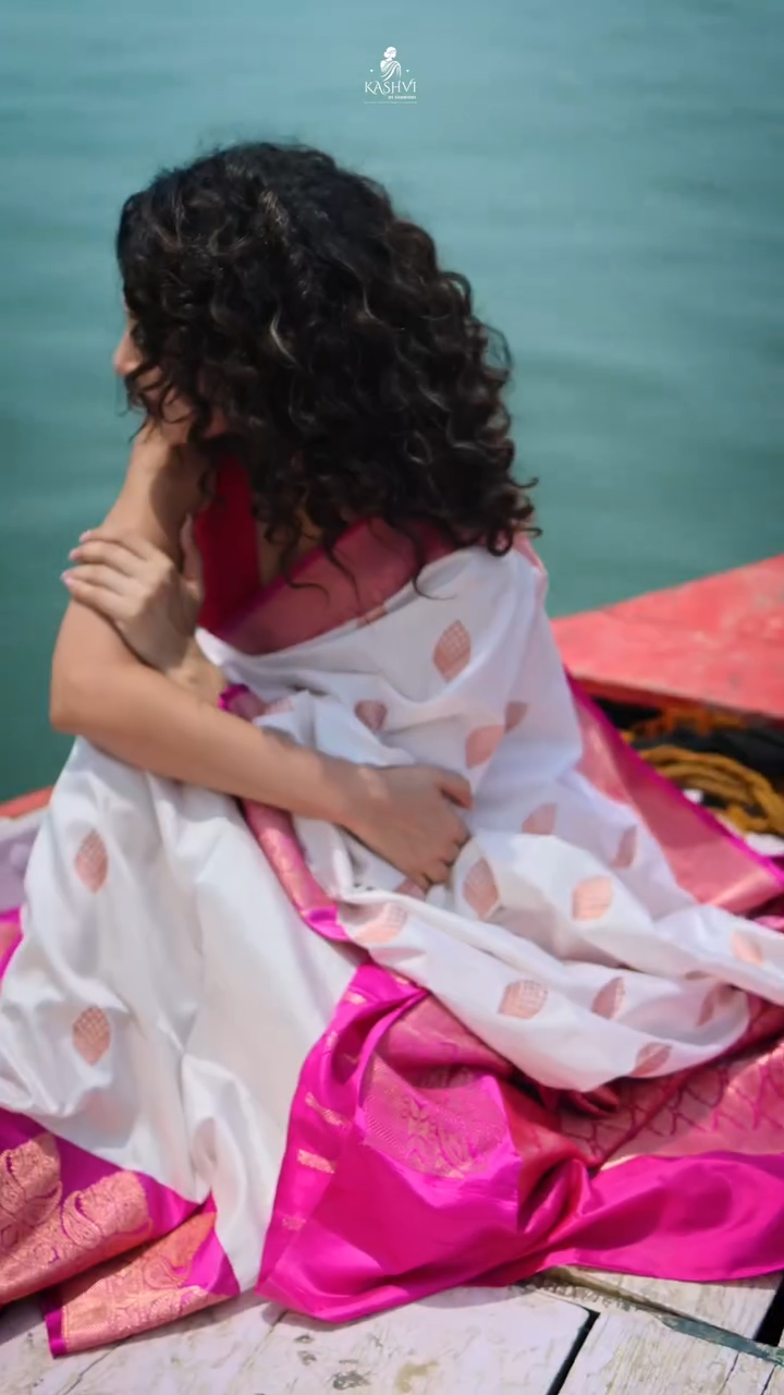 Woman in a white and pink saree sitting by a body of water.