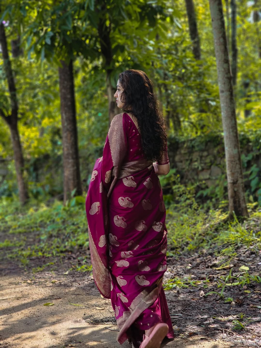 Woman in a magenta saree with gold patterns walking through a forest