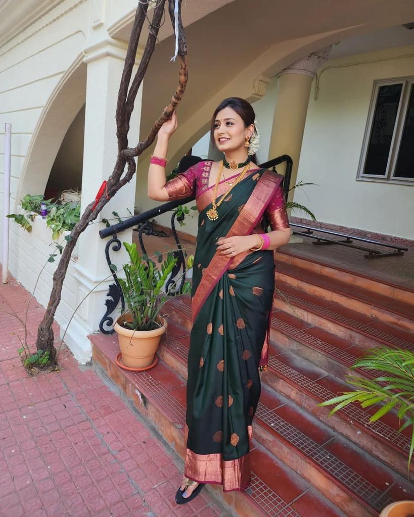 Royal purple Banarasi Silk saree with large rose gold zari paisley motifs and a heavy woven border, worn by a woman holding a small pink potli bag.