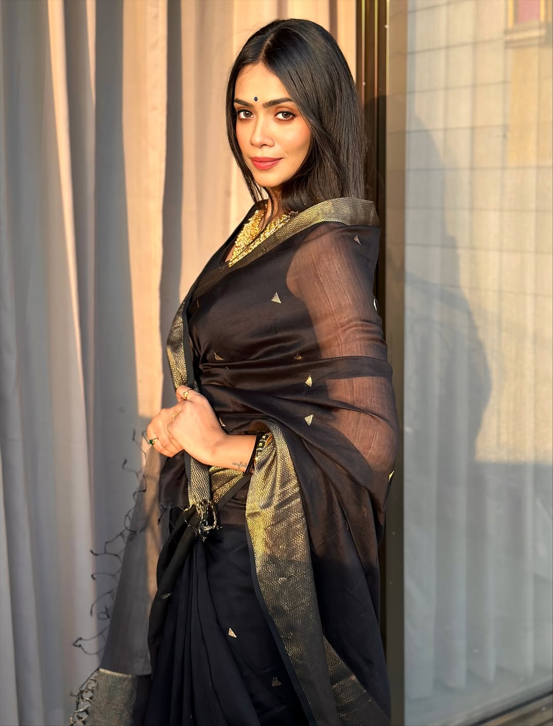 Woman in a black saree with gold border standing indoors.