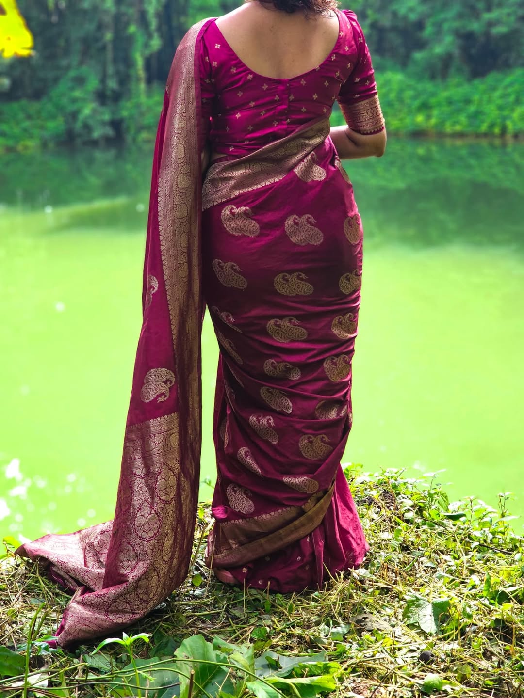 Woman wearing a pink saree with gold patterns standing by a body of water.