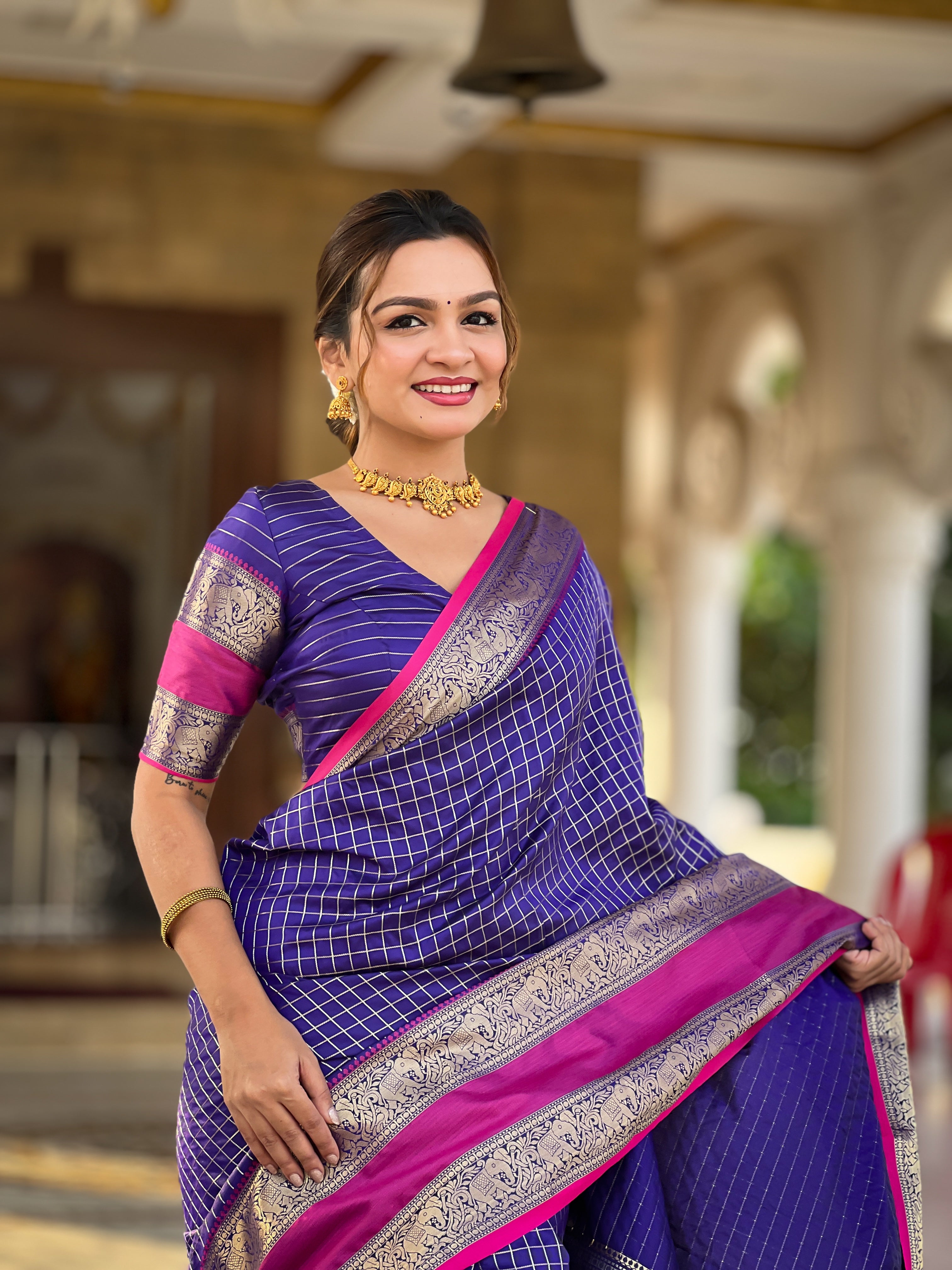 Woman wearing regal violet soft silk saree with gold zari checks, magenta contrast border, and rich pallu with tassels