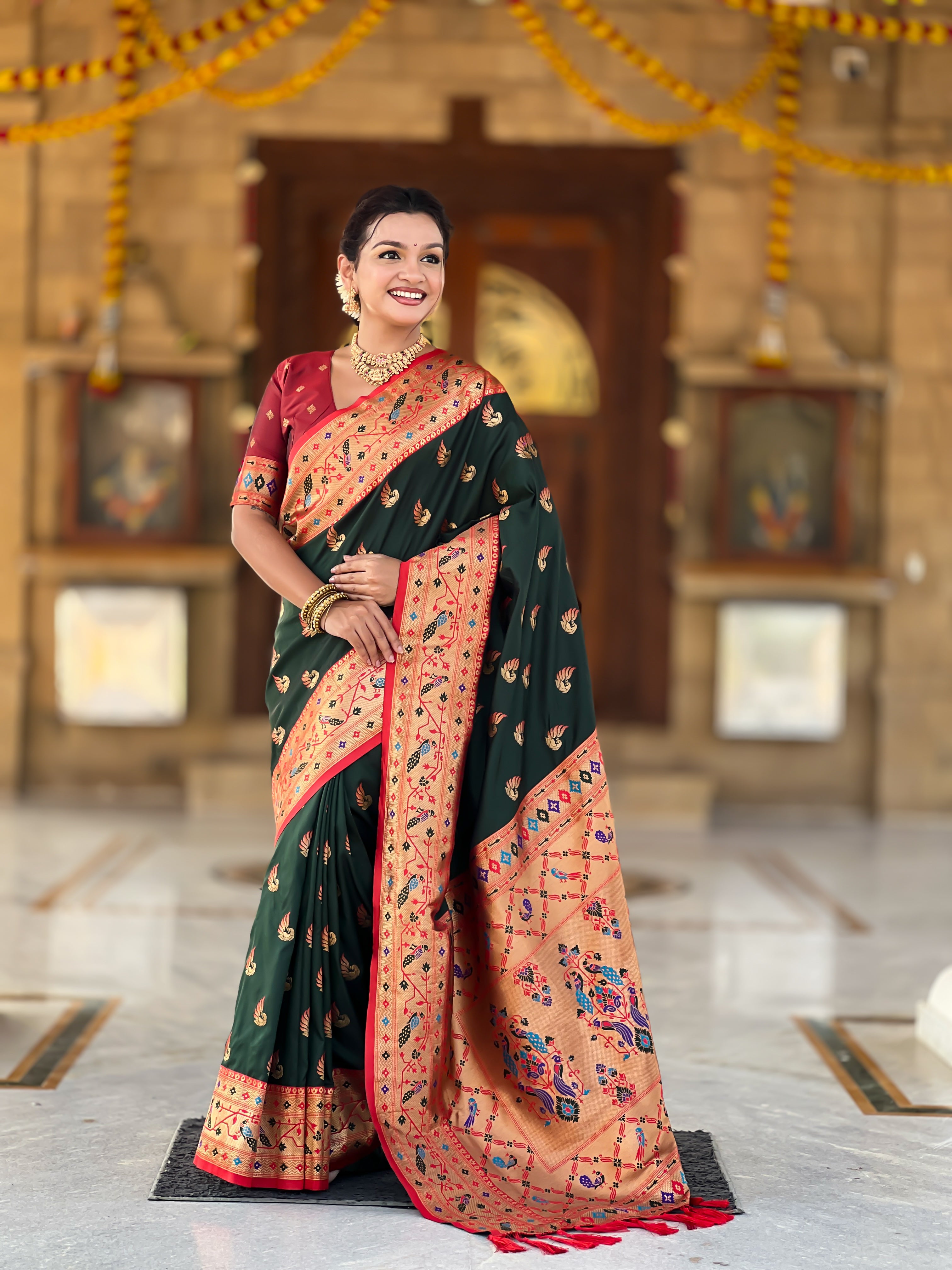 Woman wearing bottle green soft Paithani silk saree with red Meenakari border and peacock zari motifs standing in a temple setting