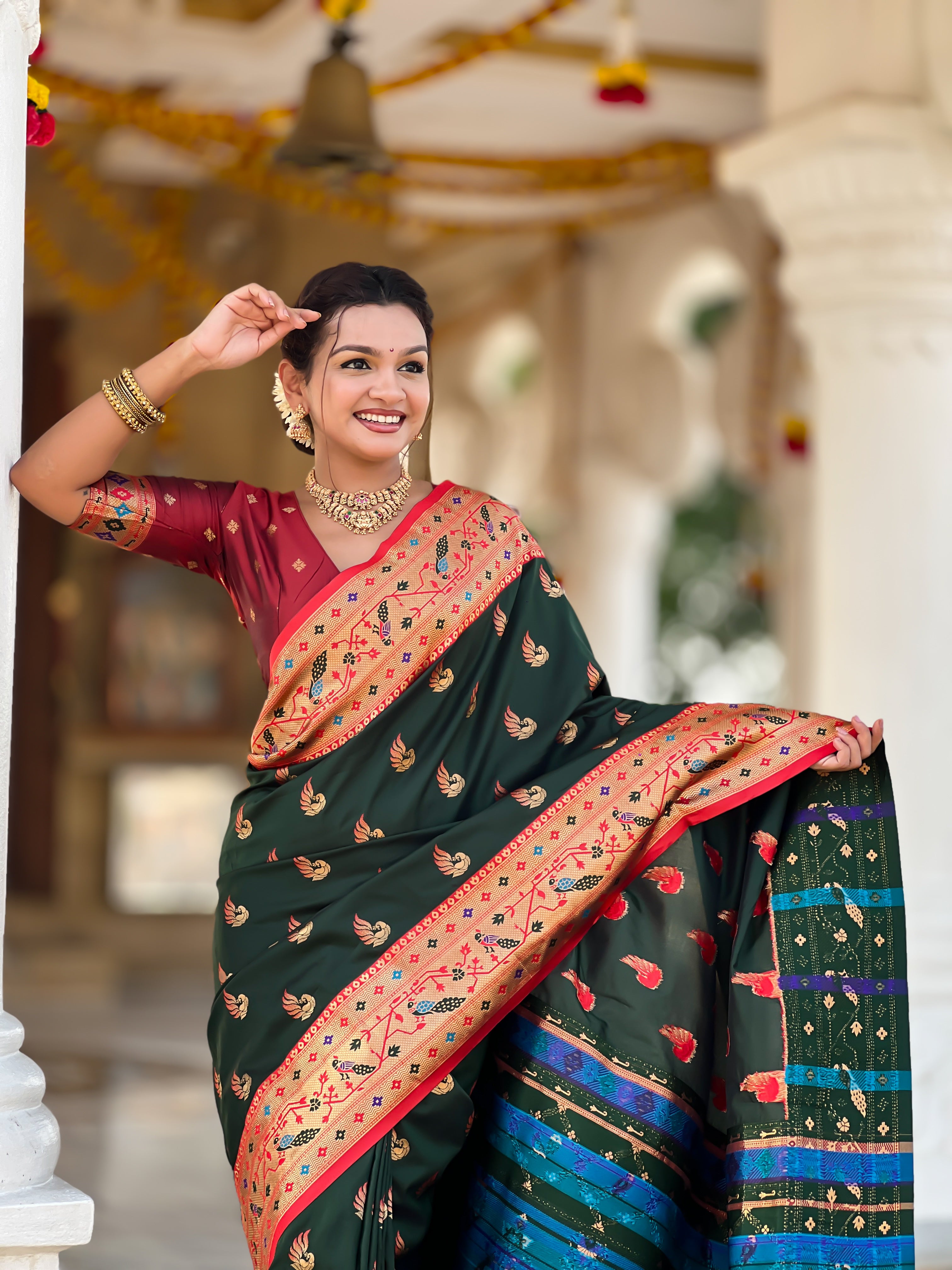 Woman wearing bottle green soft Paithani silk saree with red Meenakari border and peacock zari motifs standing in a temple setting