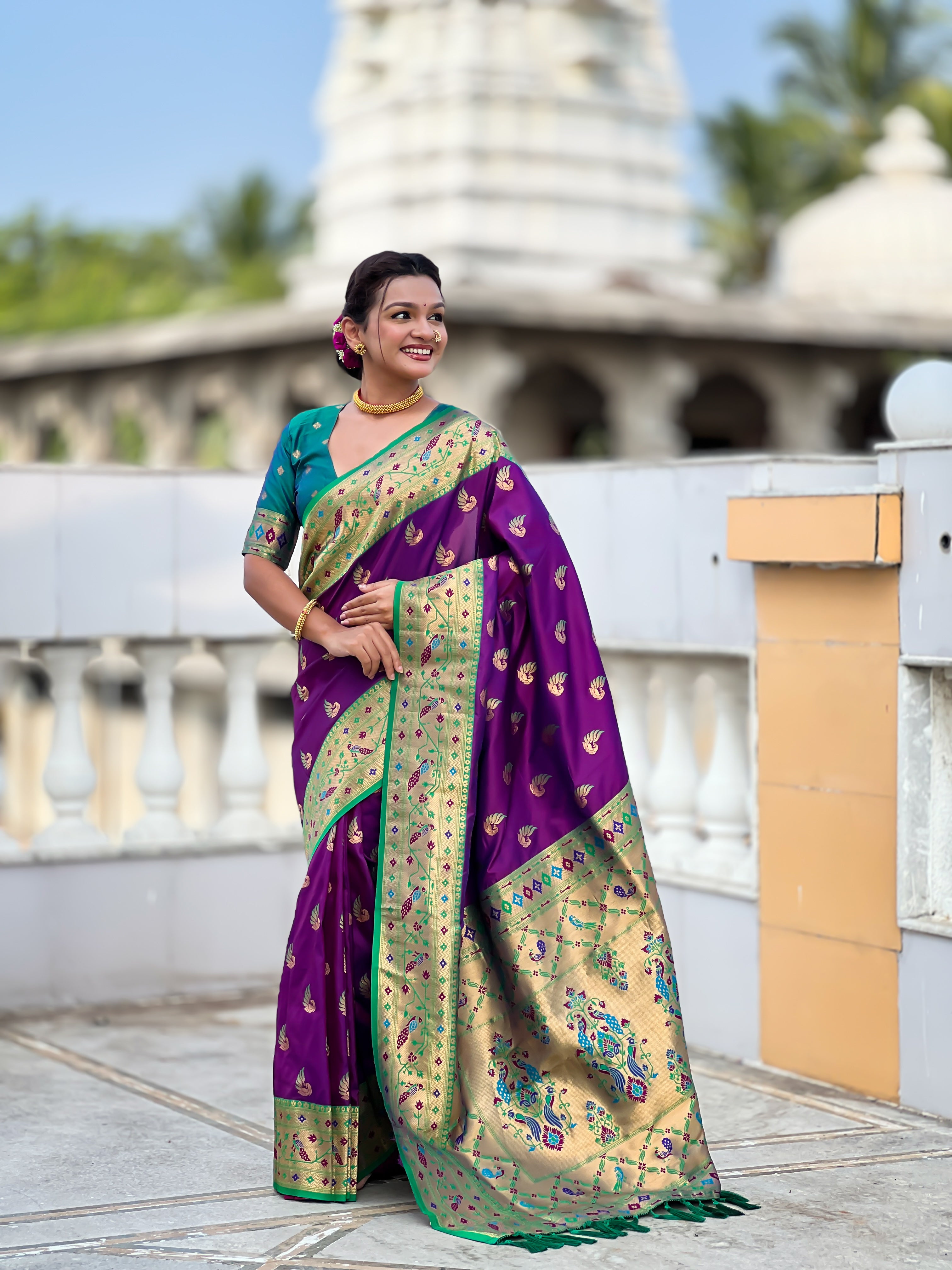 Woman wearing bottle Magenta soft Paithani silk saree with red Meenakari border and peacock zari motifs standing in a temple setting