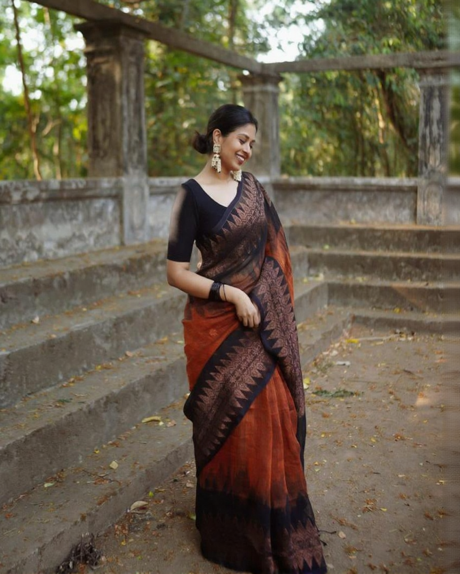 Rust orange Banarasi Silk saree with a black woven border featuring traditional motifs, worn by a smiling woman in an outdoor setting with concrete steps and greenery.